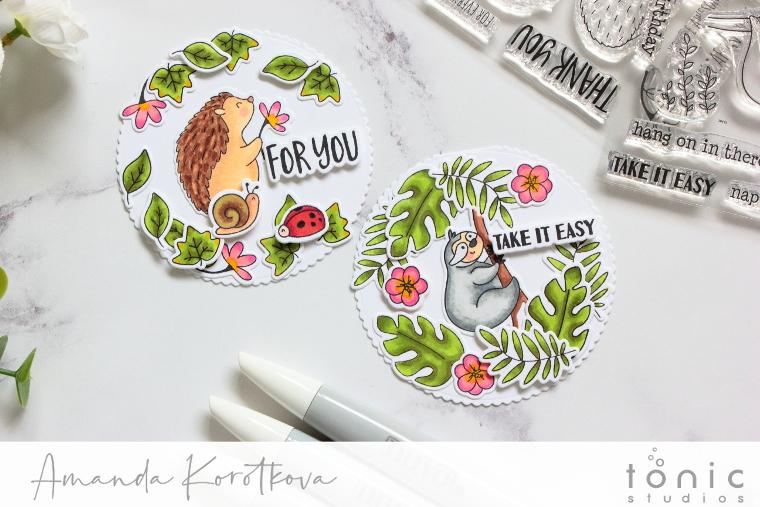 Two papercrafted badges with foliage, one had a hedgehog, snail and ladybird and the other badge has a sloth hanging onto a branch